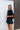 Frontal view of the City Escape Romper that features a black silky material, a rounded neckline, a flowy half sleeve, an elastic wast, a shorts bottom, side cut-outs, an open three tie back, and a flowy fit.
