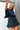 Frontal view of the City Escape Romper that features a black silky material, a rounded neckline, a flowy half sleeve, an elastic wast, a shorts bottom, side cut-outs, an open three tie back, and a flowy fit.