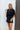 Frontal view of the City Escape Romper that features a black silky material, a rounded neckline, a flowy half sleeve, an elastic wast, a shorts bottom, side cut-outs, an open three tie back, and a flowy fit.