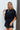 Frontal view of the City Escape Romper that features a black silky material, a rounded neckline, a flowy half sleeve, an elastic wast, a shorts bottom, side cut-outs, an open three tie back, and a flowy fit.