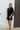 Frontal view of the City Escape Romper that features a black silky material, a rounded neckline, a flowy half sleeve, an elastic wast, a shorts bottom, side cut-outs, an open three tie back, and a flowy fit.