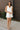 Full front view of model wearing the Everlasting Bliss Romper that has white fabric, a pleated bottom with a lettuce hem, an elastic waist, a surplice neck with a tie and snap closure, and a keyhole back.