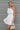 Frontal view of the Harmony Dress that features a white material, an asymmetrical neckline, a one shoulder short sleeve, a smocked waist, a flowy fit, and a mini length.