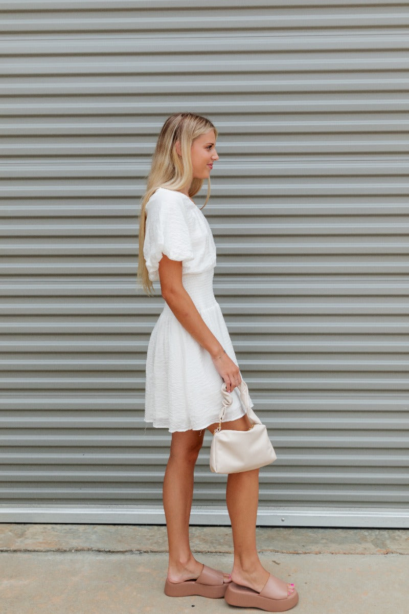 Side view of the Harmony Dress that features a white material, an asymmetrical neckline, a one shoulder short sleeve, a smocked waist, a flowy fit, and a mini length.