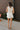 Full back view of model wearing the Everlasting Bliss Romper that has white fabric, a pleated bottom with a lettuce hem, an elastic waist, a surplice neck with a tie and snap closure, and a keyhole back.