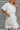 Frontal view of the Harmony Dress that features a white material, an asymmetrical neckline, a one shoulder short sleeve, a smocked waist, a flowy fit, and a mini length.