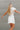 Back view of the Harmony Dress that features a white material, an asymmetrical neckline, a one shoulder short sleeve, a smocked waist, a flowy fit, and a mini length.