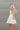 Side view of the Harmony Dress that  features a white material, an asymmetrical neckline, a one shoulder short sleeve, a smocked waist, a flowy fit, and a mini length.