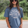 Front view of model wearing the Ford Graphic Tee that has dusty blue cotton fabric, round neckline and short sleeves. Graphic says "FORD" "MOTOR CO." "DETROIT" in white lettering. Graphic has a tire with wings in white. 