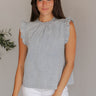 Front view of model wearing the Love Story Ruffled Top that has washed blue fabric, a round neckline with ruffle details, a back key hole with a button closure, and a sleeveless design with ruffle details.