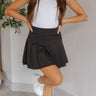 Front view of model wearing the Missing You Pleated Skirt that features black fabric with pleating, mini length, black lining, a side button, and a monochromatic side zipper with a hook closure