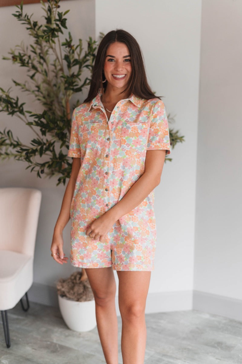 Front view of model wearing the Laguna Floral Romper that has denim fabric with an orange, pink, aqua, green and blue floral print, folded hem, two front chest pockets, side and back pockets, snap up buttons, a collared neck, and short sleeves