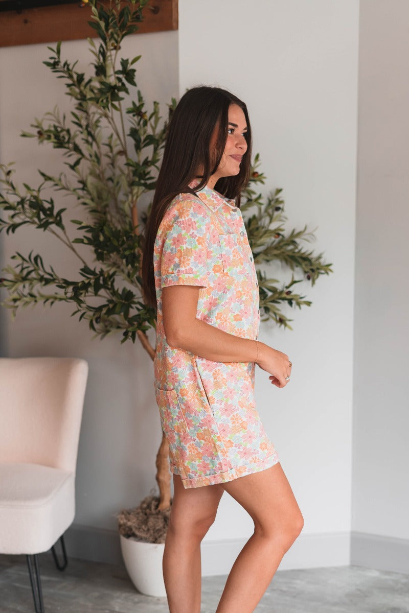 Side view of model wearing the Laguna Floral Romper that has denim fabric with an orange, pink, aqua, green and blue floral print, folded hem, two front chest pockets, side and back pockets, snap up buttons, a collared neck, and short sleeves