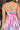 Close up back view of model wearing the In My Favor Midi Dress which features a hot pink, light pink, green, blue and lavender color block pattern, tiered ruffle details, a pink skirt lining, a square neckline, adjustable straps, a smocked back, and a mon