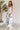 Full body view of model wearing the Garden Party Floral Dress which features white fabric with a blue floral print, midi length, white lining, a two-tiered skirt, a smocked upper with a square neckline, and ruffle straps.