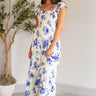 Full body view of model wearing the Garden Party Floral Dress which features white fabric with a blue floral print, midi length, white lining, a two-tiered skirt, a smocked upper with a square neckline, and ruffle straps.