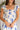 Close up view of model wearing the Garden Party Floral Dress which features white fabric with a blue floral print, midi length, white lining, a two-tiered skirt, a smocked upper with a square neckline, and ruffle straps.