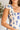 Close up view of model wearing the Garden Party Floral Dress which features white fabric with a blue floral print, midi length, white lining, a two-tiered skirt, a smocked upper with a square neckline, and ruffle straps.