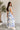 Side view of model wearing the Garden Party Floral Dress which features white fabric with a blue floral print, midi length, white lining, a two-tiered skirt, a smocked upper with a square neckline, and ruffle straps.