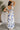 Back view of model wearing the Garden Party Floral Dress which features white fabric with a blue floral print, midi length, white lining, a two-tiered skirt, a smocked upper with a square neckline, and ruffle straps.