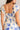 Close up back view of model wearing the Garden Party Floral Dress which features white fabric with a blue floral print, midi length, white lining, a two-tiered skirt, a smocked upper with a square neckline, and ruffle straps.