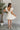 Full body back view of model wearing the One More Time Dress that has cream sheer fabric, a ruffled stripe design, a bubble hem, cream lining, a plunge neckline, ruffle straps, an open back and a monochromatic back zipper with a hook closure