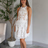 Front view of model wearing the Everlasting Love Mini Dress which features white sheer fabric with white floral embroidery, white tulle lining, tan lining, mini length, a halter neckline with a tie closure, and an open back with button closures.