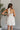 Back view of model wearing the Everlasting Love Mini Dress which features white sheer fabric with white floral embroidery, white tulle lining, tan lining, mini length, a halter neckline with a tie closure, and an open back with button closures.