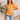 Front view of model wearing the Serena Floral Top which features orange fabric with a teal, mustard, tan and white floral print, a peplum body, a square neckline, short puff sleeves, and a smocked back.