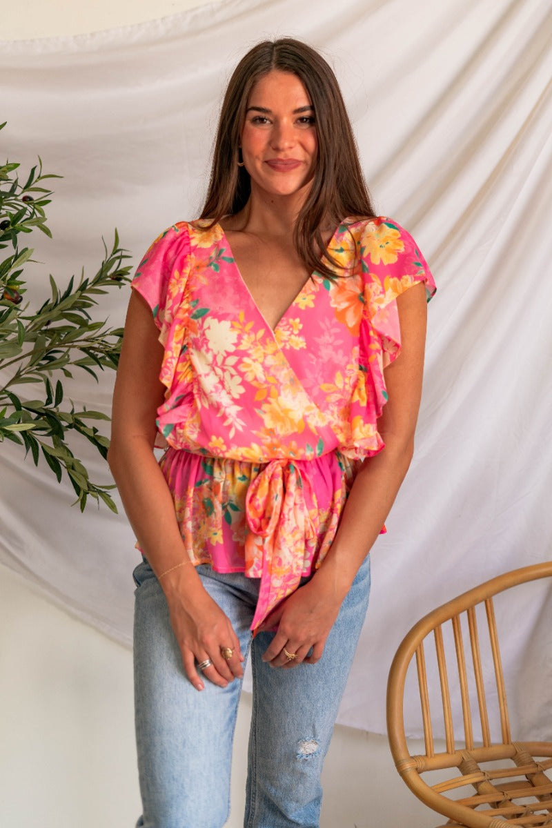 Front view of model wearing the Luna Floral Top which features pink fabric with yellow, green, orange and white floral print, an elastic waistband, a tie around the waist, a surplice neckline with a clasp closure, and short sleeves with ruffle details.