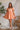 Full body view of model wearing the Into The Sunset Dress which features peach fabric with peach lining, square neckline, short puff sleeves, a smocked back and a monochromatic side zipper with a hook closure.