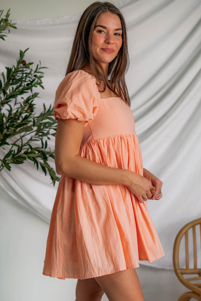 Side view of model wearing the Into The Sunset Dress which features peach fabric with peach lining, square neckline, short puff sleeves, a smocked back and a monochromatic side zipper with a hook closure.