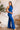 Side view of model wearing the Get On My Level Jumpsuit that has royal blue fabric, a front key hole , a halter neck with button closures, a sleeveless design, an open back, a back zipper with a hook closure, and flare pant legs.