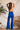 Back view of model wearing the Get On My Level Jumpsuit that has royal blue fabric, a front key hole , a halter neck with button closures, a sleeveless design, an open back, a back zipper with a hook closure, and flare pant legs.