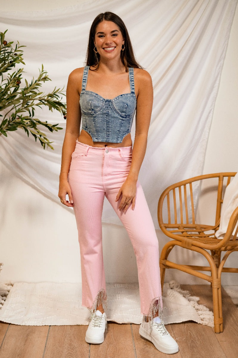Full body front view of model wearing the Just Dance Rhinestone Fringe Jeans that have light pink denim fabric, a front zipper, belt loops, a frayed waist, pockets, and a high-low hem with rhinestone fringe details