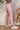 Side view of model wearing the Just Dance Rhinestone Fringe Jeans that have light pink denim fabric, a front zipper, belt loops, a frayed waist, pockets, and a high-low hem with rhinestone fringe details