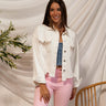 Front view of model wearing the Roxanne Rhinestone Fringe Jacket that has white denim fabric, a frayed hem, silver buttons, a collar, two front chest pockets with rhinestone fringe details, and long sleeves