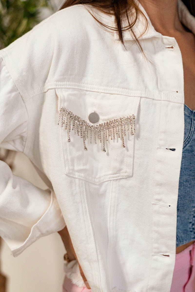 Close up view of model wearing the Roxanne Rhinestone Fringe Jacket that has white denim fabric, a frayed hem, silver buttons, a collar, two front chest pockets with rhinestone fringe details, and long sleeves