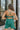 Back view of the Something Like That Dress that features a teal green satin material, a cowl neckline with cups at the bust, a sleeveless design with thin adjustable straps, a waist cut-out, an elastic waistline, and a criss-cross strap back