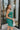 Side view of the Something Like That Dress that features a teal green satin material, a cowl neckline with cups at the bust, a sleeveless design with thin adjustable straps, a waist cut-out, an elastic waistline, and a criss-cross strap back