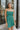 Frontal view of the Something Like That Dress that features a teal green satin material, a cowl neckline with cups at the bust, a sleeveless design with thin adjustable straps, a waist cut-out, an elastic waistline, and a criss-cross strap back