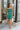 Frontal view of the Something Like That Dress that features a teal green satin material, a cowl neckline with cups at the bust, a sleeveless design with thin adjustable straps, a waist cut-out, an elastic waistline, and a criss-cross strap back