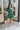 Frontal view of the Juniper Romper that features a green material, an asymmetrical neckline, a one shoulder short puffy sleeve, pockets, a waist tie detail, and a hidden side zipper.