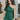 Frontal view of the Juniper Romper that features a green material, an asymmetrical neckline, a one shoulder short puffy sleeve, pockets, a waist tie detail, and a hidden side zipper.