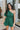 Frontal view of the Juniper Romper that features a green material, an asymmetrical neckline, a one shoulder short puffy sleeve, pockets, a waist tie detail, and a hidden side zipper.