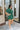 Side view of the Juniper Romper that features a green material, an asymmetrical neckline, a one shoulder short puffy sleeve, pockets, a waist tie detail, and a hidden side zipper.