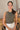 Front view of model wearing the Call It Even Bodysuit, that features stretchy olive fabric, a round neckline, and a thong bottom with hook closures. Worn with white jeans.