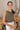 Front view of model wearing the Call It Even Bodysuit, that features stretchy olive fabric, a round neckline, and a thong bottom with hook closures. Worn with white jeans.
