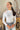 Front view of model wearing the On The Daily Bodysuit, that has white textured fabric, a high neck with lettuce trim, long sleeves with lettuce trim, and a thong bottom with hook closures. Worn with jeans.