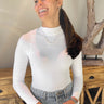 Front view of model wearing the On The Daily Bodysuit, that has white textured fabric, a high neck with lettuce trim, long sleeves with lettuce trim, and a thong bottom with hook closures. Worn with jeans.